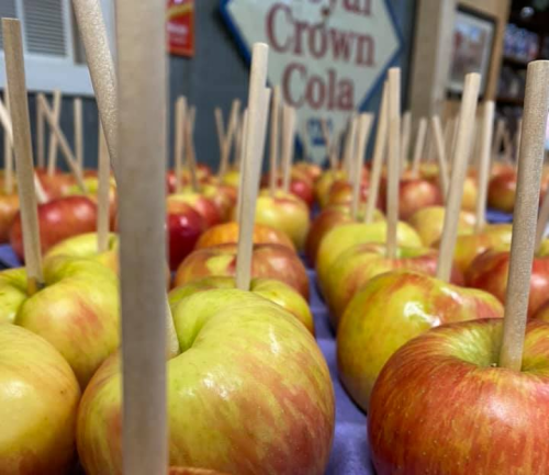 A close-up of apples with wooden sticks inserted, arranged on a purple surface, with a vintage soda sign in the background.
