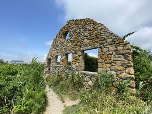 A weathered stone ruin with arched windows, surrounded by tall grass and greenery under a blue sky.