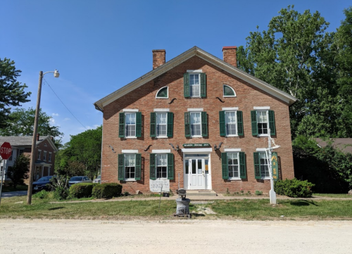 Historic brick building with green shutters, white door, and a sign in front, surrounded by greenery and a clear blue sky.