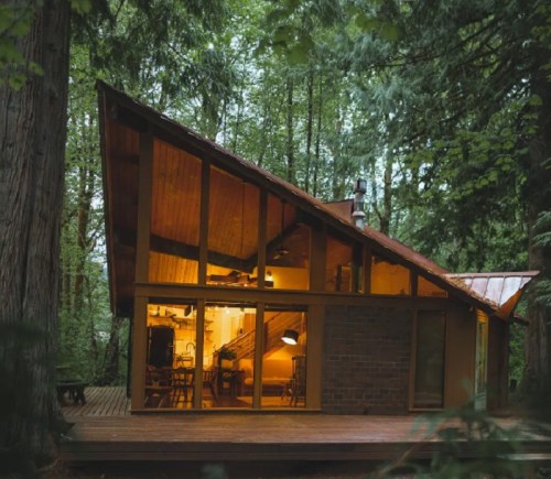 A modern cabin with a sloped roof nestled among tall trees, illuminated warmly from within.