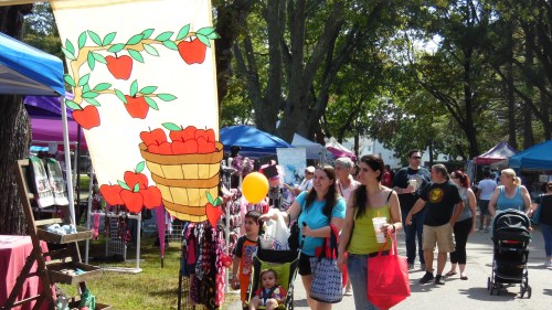 A lively outdoor market scene with colorful tents, shoppers, and a banner featuring a basket of apples.