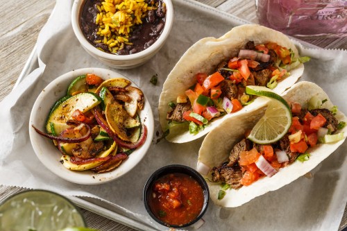 Two tacos filled with beef, tomatoes, and lime, served with black beans, rice, and sautéed vegetables on a tray.