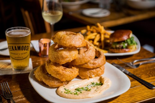A plate of stacked onion rings with dipping sauce, a burger, fries, and a glass of beer on a wooden table.