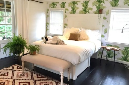 Cozy bedroom with a large bed, tropical wallpaper, a bench, and a rug, surrounded by natural light from windows.