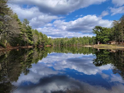 A serene lake reflecting trees and clouds under a blue sky, surrounded by lush greenery.