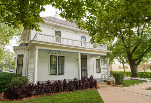 A charming white two-story house with a balcony, surrounded by green trees and colorful plants.