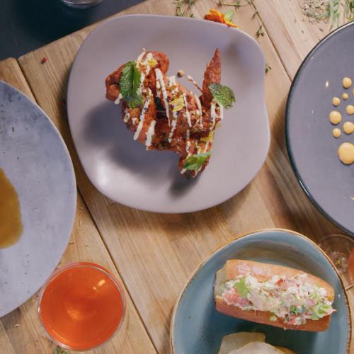 A plate of crispy fried chicken topped with herbs and drizzled with sauce, surrounded by various dishes and a drink.