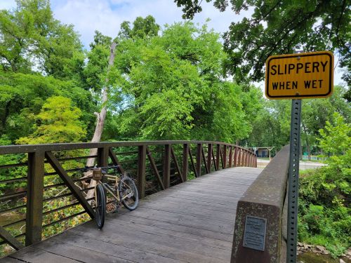 A wooden bridge surrounded by lush greenery, with a bike parked and a warning sign that says "Slippery When Wet."