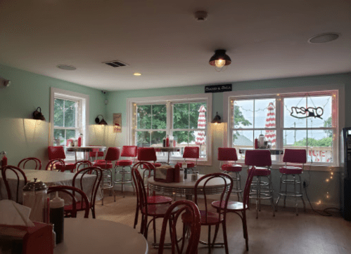 A cozy diner interior with red chairs, tables, and large windows featuring striped umbrellas outside.