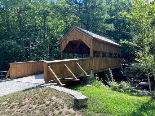 A wooden covered bridge spans a small stream, surrounded by lush greenery and trees.
