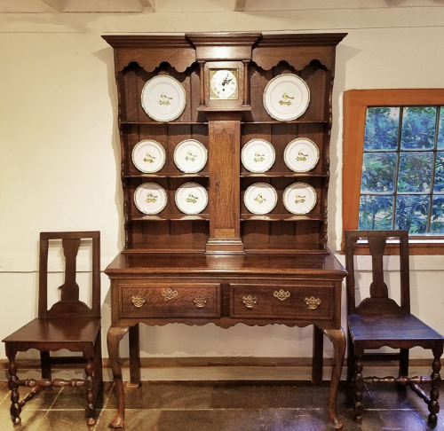 A wooden hutch with decorative plates on display, flanked by two matching chairs in a well-lit room.