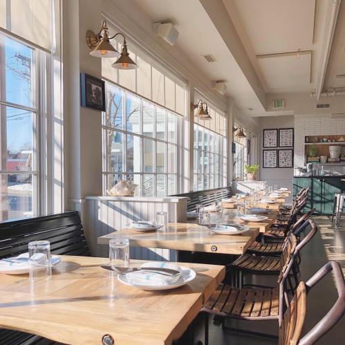 Bright restaurant interior with wooden tables, chairs, and large windows, creating a warm and inviting atmosphere.