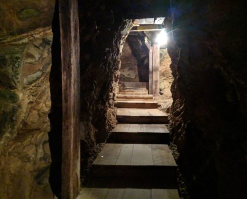 Dimly lit underground passage with wooden steps and stone walls, leading deeper into a mine.