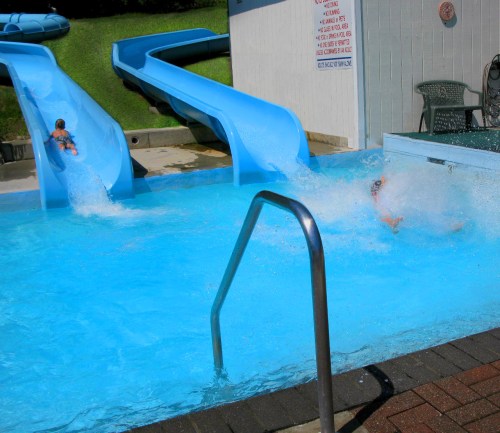 Two children slide down blue water slides into a pool, creating splashes in the bright blue water.