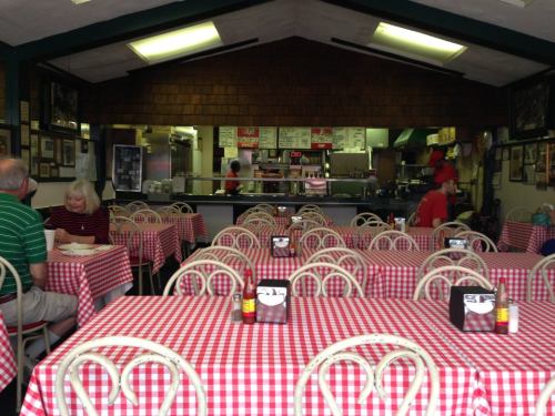 Cozy diner interior with red and white checkered tables, empty seats, and a visible kitchen in the background.
