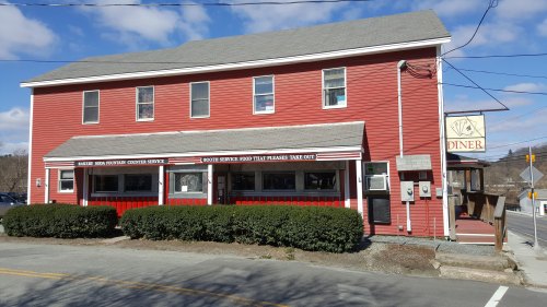 A red diner building with large windows and a sign, surrounded by greenery and a road in front.
