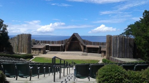 An outdoor theater with wooden structures, surrounded by greenery and overlooking a calm body of water under a blue sky.