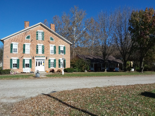 A large brick building with green shutters, surrounded by trees and a gravel road on a clear day.