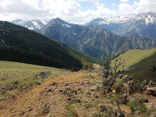 A scenic mountain landscape with rugged terrain, lush greenery, and snow-capped peaks under a partly cloudy sky.