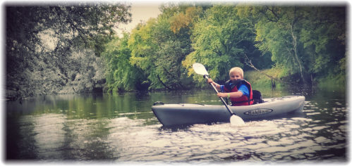 A child paddles a kayak on a calm river surrounded by lush green trees.