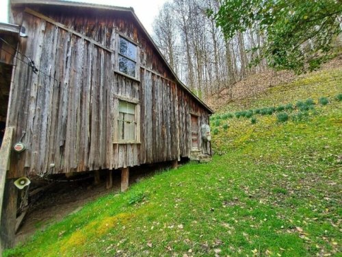A rustic wooden barn stands on a grassy slope, surrounded by trees and a hillside of greenery.