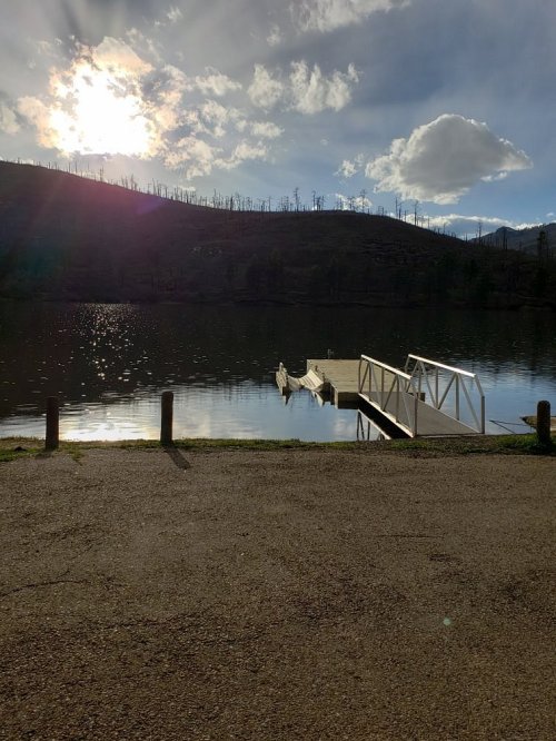 A serene lake scene with a dock, surrounded by hills and a cloudy sky, reflecting sunlight on the water's surface.