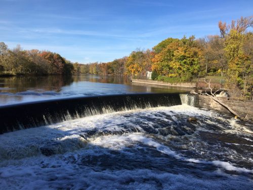 A serene river scene with a waterfall, surrounded by trees displaying autumn colors under a clear blue sky.