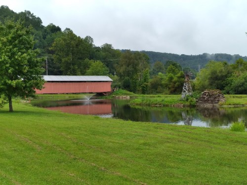 A red covered bridge spans a calm pond, surrounded by lush greenery and distant hills under a cloudy sky.