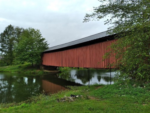 A red covered bridge spans a calm river, surrounded by green trees and grass under a cloudy sky.
