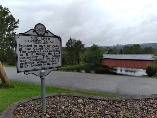 A historical marker for Mud River Covered Bridge, with the bridge visible in the background and a serene landscape.