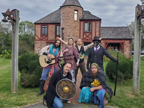 A group of six people in costumes poses outside a brick house, with one holding a guitar and another a shield.