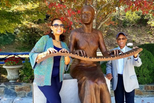 Two people pose beside a bronze statue of a seated woman, surrounded by colorful autumn foliage.