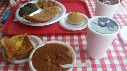 A tray with fried fish, greens, apples, a roll, toast, and a bowl of beans, alongside a large drink on a checkered tablecloth.