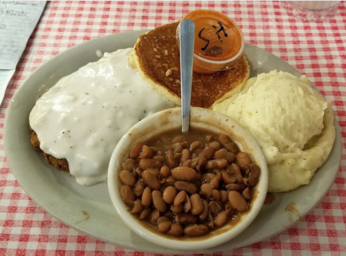 A plate with chicken fried steak topped with gravy, pancakes, mashed potatoes, and a side of pinto beans.