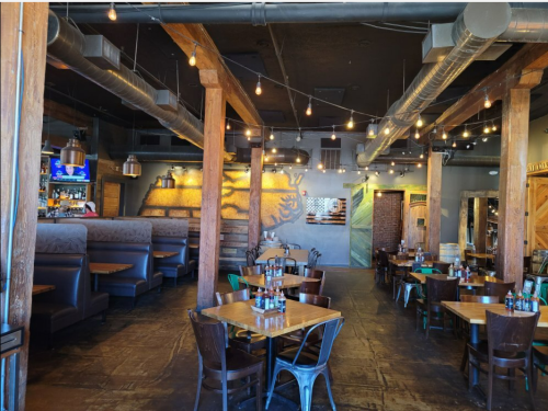 Interior of a restaurant featuring wooden beams, booths, tables, and warm lighting with a rustic decor.