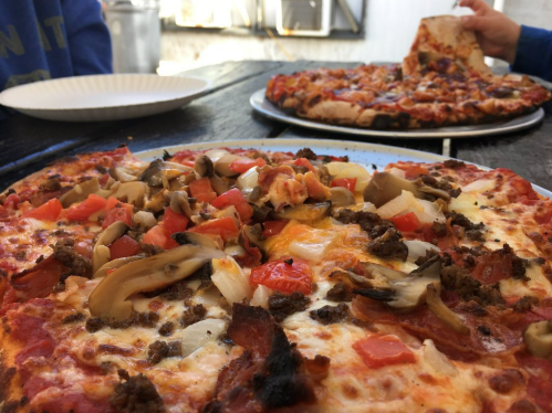Two pizzas on a wooden table, one topped with mushrooms, tomatoes, and ground meat, the other partially eaten.