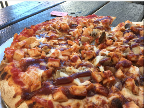 A close-up of a pizza topped with diced chicken, barbecue sauce, and mushrooms on a wooden table.
