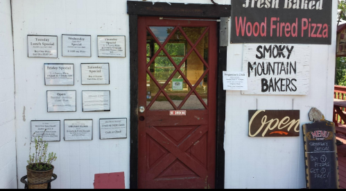 A rustic entrance with a red door, featuring a sign for wood-fired pizza and daily specials on the wall.