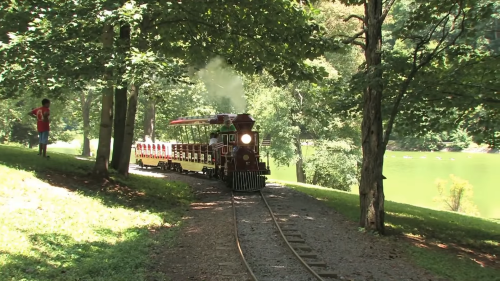A vintage train travels along a scenic path by a lake, surrounded by lush green trees. A person walks nearby.