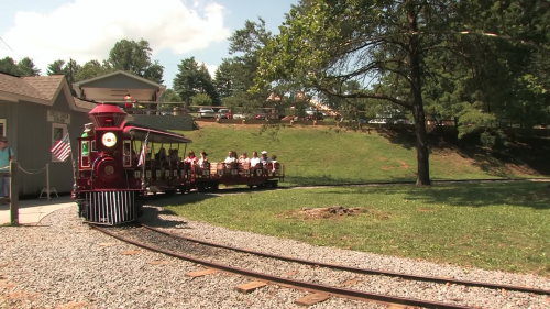 A vintage train with passengers travels along a gravel track in a scenic outdoor setting.