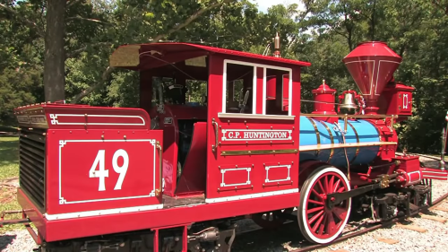 A vintage red steam locomotive with blue accents, labeled "C.P. Huntington," set in a green outdoor environment.