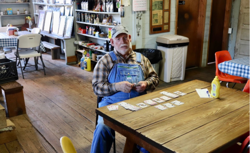 An older man in overalls sits at a table playing cards in a rustic room with wooden floors and shelves.
