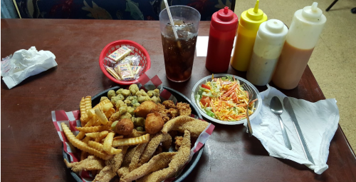A plate of fried fish, crinkle-cut fries, hushpuppies, okra, and a side salad, with condiments and a drink.