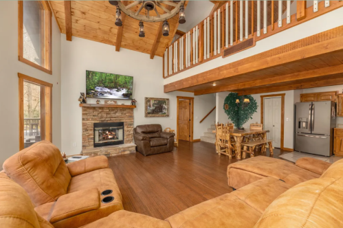 Cozy living room with a stone fireplace, wooden beams, and a dining area, featuring large windows and comfortable seating.