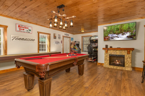 A cozy game room featuring a pool table, fireplace, TV, and rustic decor with wooden walls and a warm atmosphere.