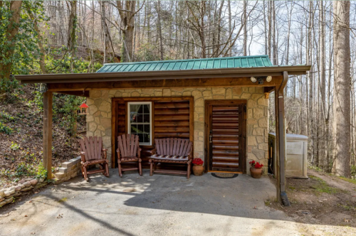 A cozy stone cabin with a green metal roof, surrounded by trees, featuring wooden chairs on the porch.