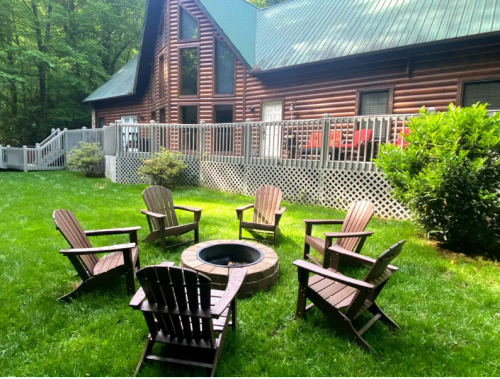 A cozy outdoor seating area with wooden chairs around a fire pit, set against a log cabin and green lawn.