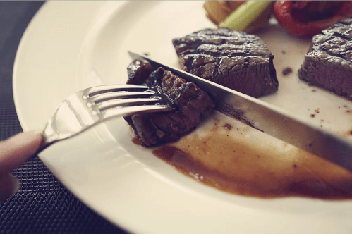 A close-up of a fork cutting into a piece of steak on a plate, accompanied by vegetables and sauce.