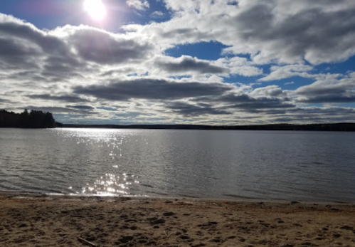 A serene lake scene with sparkling water, sandy shore, and a cloudy sky illuminated by the sun.