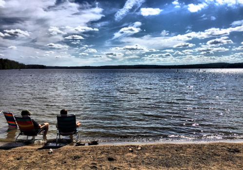 Two people sit in chairs by a calm lake, enjoying a sunny day with clouds in the sky and water reflecting the light.
